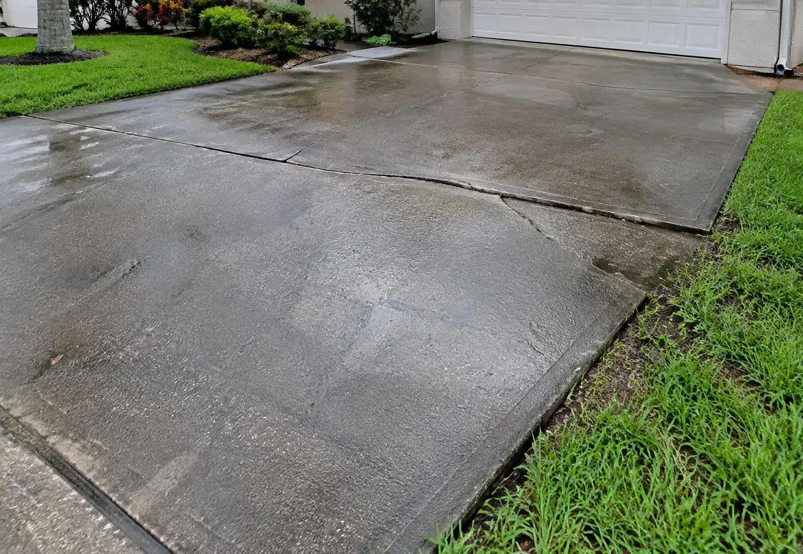 Older concrete driveway with long cracks and a slightly uneven section after rain
